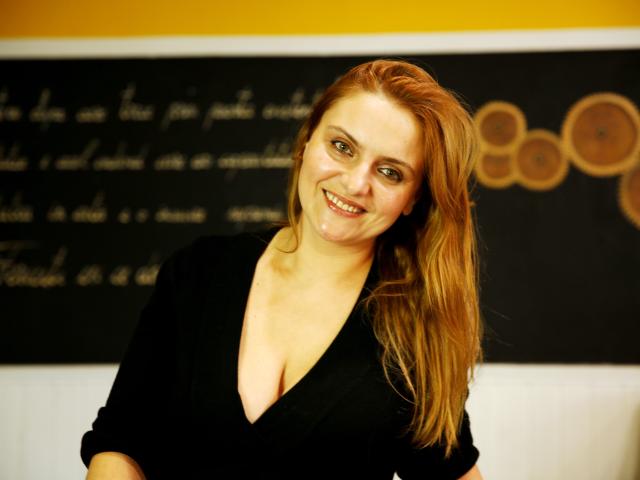 Oliviaaa, avec des cheveux roux, souriante, portant un haut noir en V qui met en valeur sa poitrine généreuse, se tient devant un tableau noir. Son visage est légèrement maculé de matière blanche..