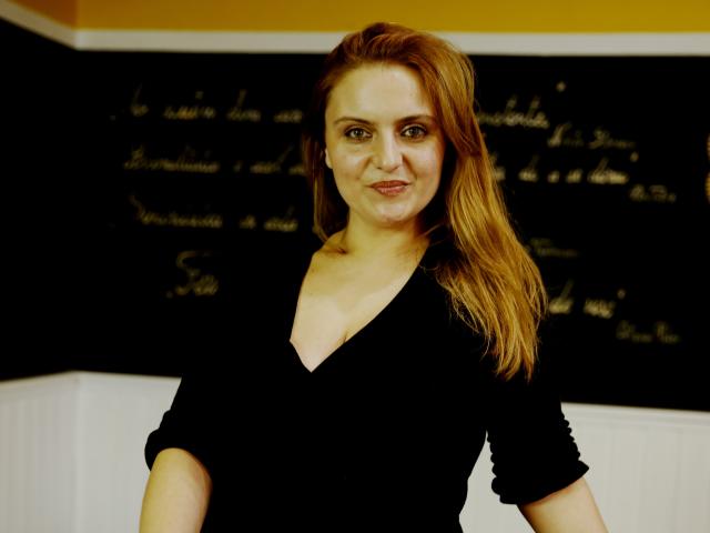 Oliviaaa with red hair, wearing a black V-neck top, smiling at the camera, showing her boobs slightly, standing in front of a black chalkboard.