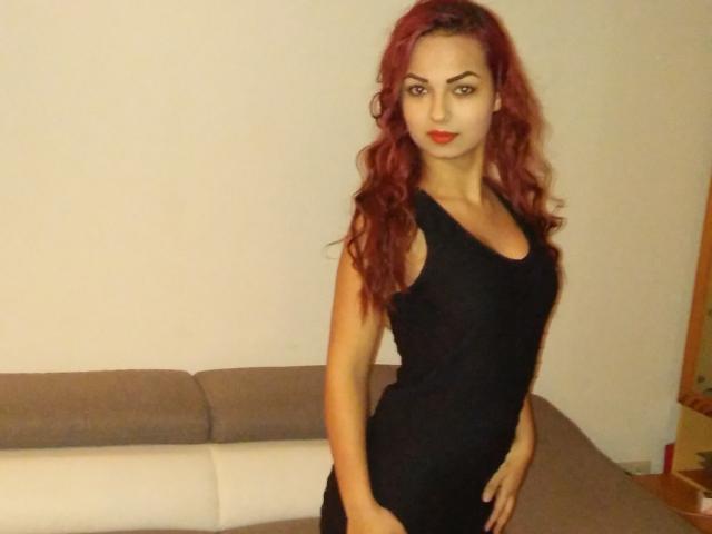 EmmaHotX69 mit roten Haaren, trägt ein enges schwarzes Kleid, zeigt ihr Gesäß, steht in einem Raum mit einem grauen und weißen Sofa und schaut in die Kamera..