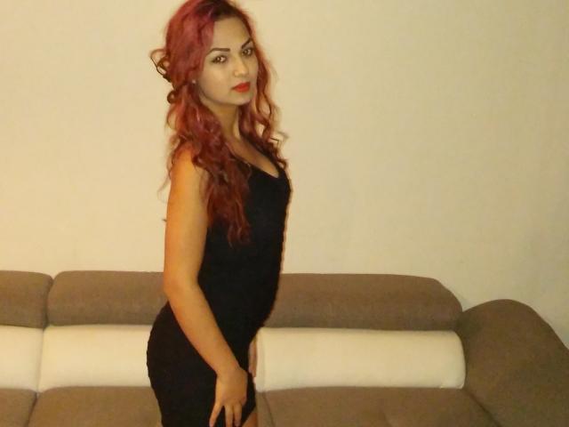 EmmaHotX69, avec des cheveux roux bouclés, une robe noire et du rouge à lèvres rouge, se tient devant un canapé beige, ses fesses légèrement relevées, les mains sur ses cuisses, et regarde l'appareil photo..