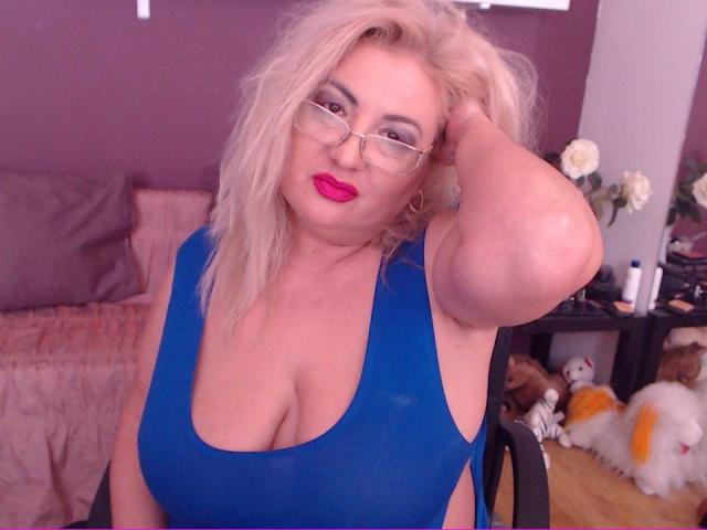 TresSexyFlorence, con i capelli biondi, gli occhiali e il rossetto rosso, indossa una maglietta blu, scollata, che mette in mostra il suo seno grande e formoso, e si tocca la testa. È seduta su una sedia..