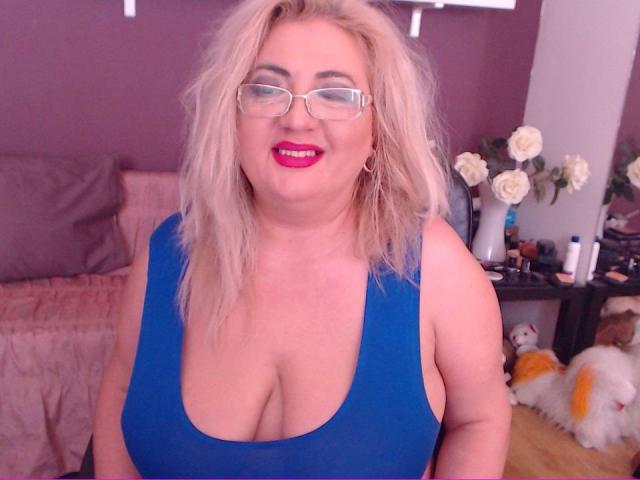 TresSexyFlorence with blonde hair, glasses, and red lipstick, wearing a blue tank top that shows her big, chubby tits. She's smiling and has a pink bedroom in the background.