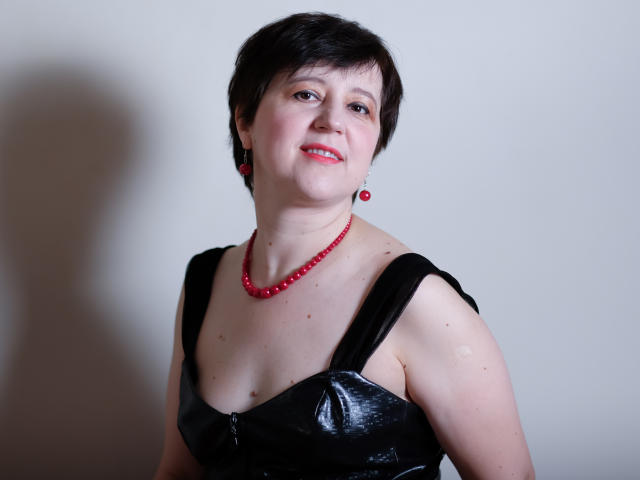 ChristaRose, avec ses cheveux noirs courts, ses boucles d'oreilles rouges, son collier de perles rouges et sa robe noire brillante, dévoilant ses petites seins et souriant légèrement. Une légère ombre sur la gauche..