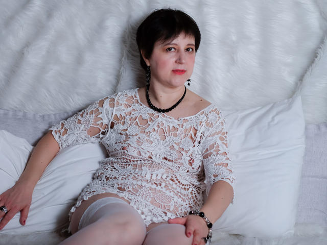 ChristaRose, short dark hair, white lace dress, visible pussy through dress, white thigh-highs, black necklace, sitting on white pillows, looking at camera, red lipstick, gentle smile.
