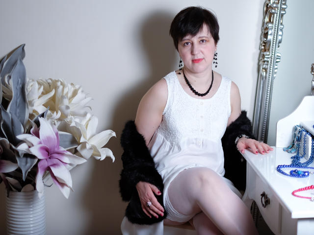 ChristaRose, con i capelli neri corti, indossa un abito bianco in pizzo, un cappotto di pelliccia nera, calze bianche, una collana di perline nere e orecchini, seduta su una sedia, con le gambe incrociate e un sorriso..