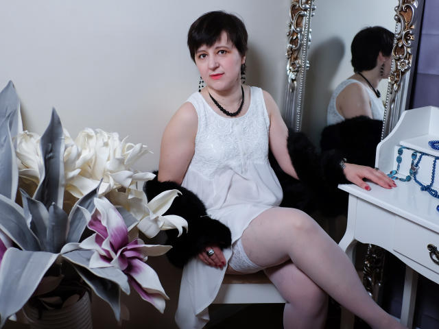 ChristaRose, short black hair, white dress, lace top, black fur, white stockings, sitting, crossed legs, black necklace, mirror reflection, white flowers, potted plant, white dresser, blue beads.