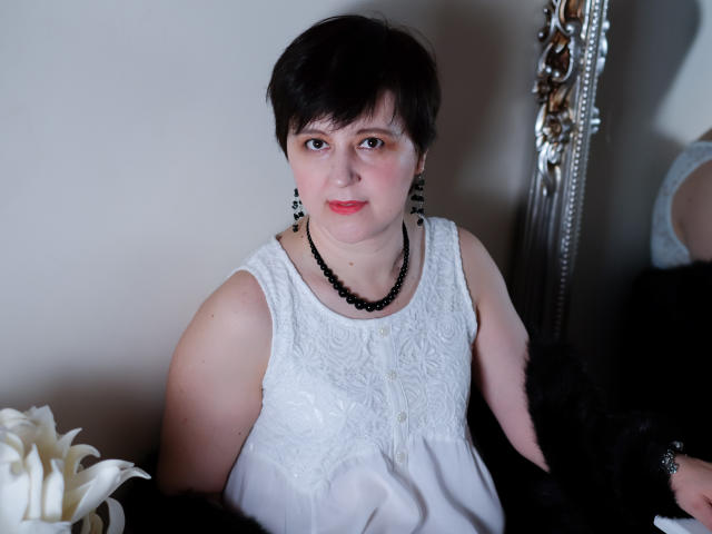 ChristaRose has short black hair, red lipstick, and is wearing a white lace dress with black bead necklace and earrings. She has small tits and a black fur shawl.