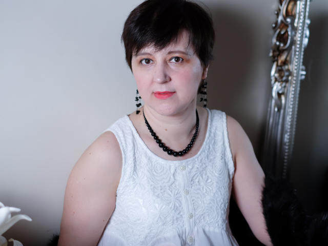 ChristaRose has short black hair, fair skin, small tits, wearing a white lace dress, black beaded necklace, and earrings, with a neutral expression and pink lipstick.
