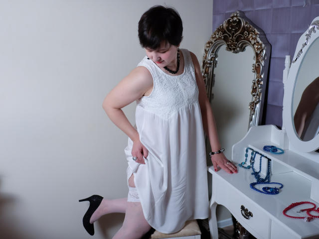 ChristaRose with short black hair, white lace dress, black heels, and white garter, kneeling, showing her pussy, next to a white vanity with jewelry.