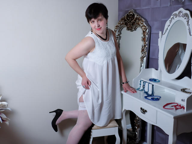 ChristaRose, short black hair, white dress, lifting it to show white lace thigh-highs and black high heels, standing in front of a white vanity with ornate mirror.