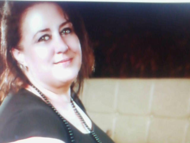 Una mujer atractiva, de buen tamaño, con tetas medianas, cabello castaño, camisa negra y collar, sonriendo a la cámara, piel clara, fondo oscuro..