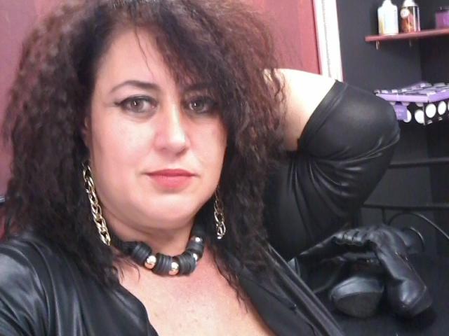 Una mujer atractiva, voluptuosa, con cabello castaño rizado, vestida con un conjunto de cuero negro, aretes de cadena, un collar negro, guantes negros y lápiz labial rojo, posando con un brazo detrás de la cabeza..