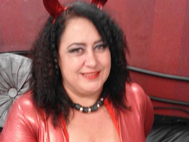 Una donna attraente, formosa, con capelli neri ricci, corna da diavolo rosse, una maglietta rossa lucida, una collana di perline nere, che sorride, con rossetto rosso, seno di media grandezza, pelle chiara, in una camera da letto..