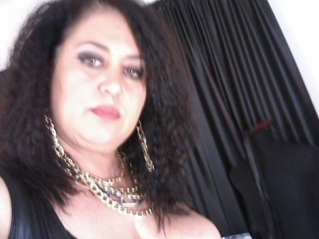 SeductiveBustyBabe with dark curly hair, heavy makeup, gold chain necklace, black leather top, showing her large tits and pink lipstick, in a slightly blurred selfie.