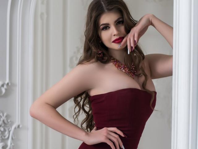ValeriMair with long brown hair, red lipstick, and a strapless maroon dress, showing off her tits and ass, wearing a red necklace, and resting her hand on her face.