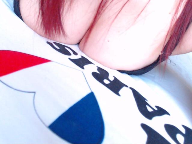 DouceAnna's tits are pressed against a white shirt with red, blue, and black patterns. Her red hair is partially visible. The shirt's text is not shown.