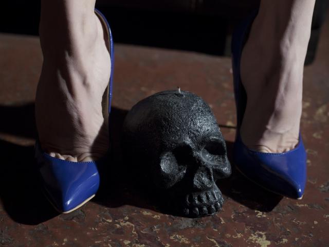 ClaraDomme's bleke benen, gekleed in glanzende blauwe schoenen, staan bovenop een zwarte schedel op een ruw, bruin oppervlak. Haar tenen zijn gespreid, waardoor de gedetailleerde, getextureerde tanden van de schedel zichtbaar zijn..
