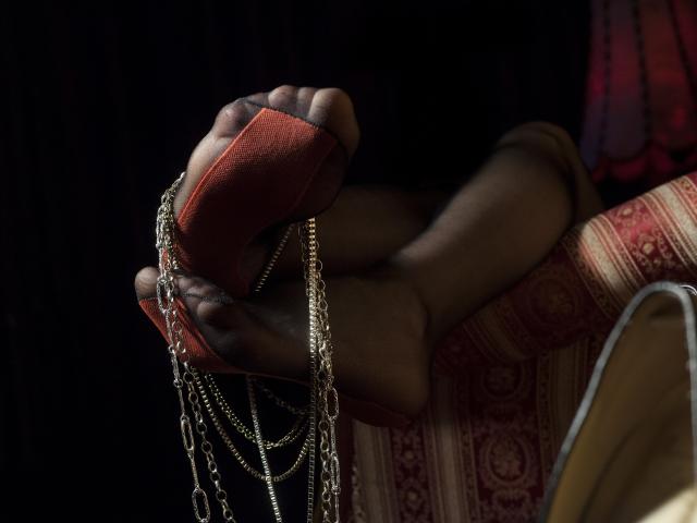 ClaraDomme's dark brown foot holds a red sandal, draped with gold chains, resting on a red-patterned, yellow-edged chair. Her toes are visible, with a relaxed posture.