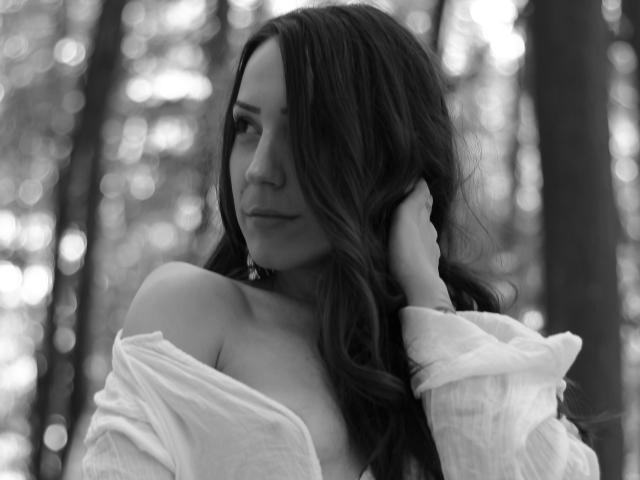 Black-and-white photo of RubinRossey with long, wavy hair covering one eye, wearing an off-shoulder white shirt, hand in hair, looking to the side, soft expression.