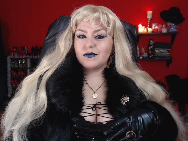 KaryQueen con i capelli biondi, un cappotto di pelliccia nera, una camicetta con lacci che lascia intravedere il seno, rossetto blu, guanti neri, una collana d'oro e una spilla a forma di fiore, in una stanza rossa..