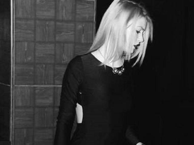 Schwarz-weißes Foto von BlossomPussy, einer blonden Frau mit schulterlangem Haar, die ein schwarzes Oberteil mit langen Ärmeln, eine Halskette und ein Kleid mit offenem Rücken trägt und mit leicht geöffnetem Mund steht..