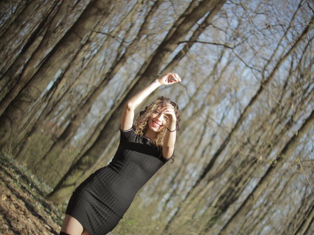 PoxyVibe, with curly blonde hair, wearing a tight black dress, has her arms raised, smiling in a sunlit forest. Her tits are small, and her ass is cute.