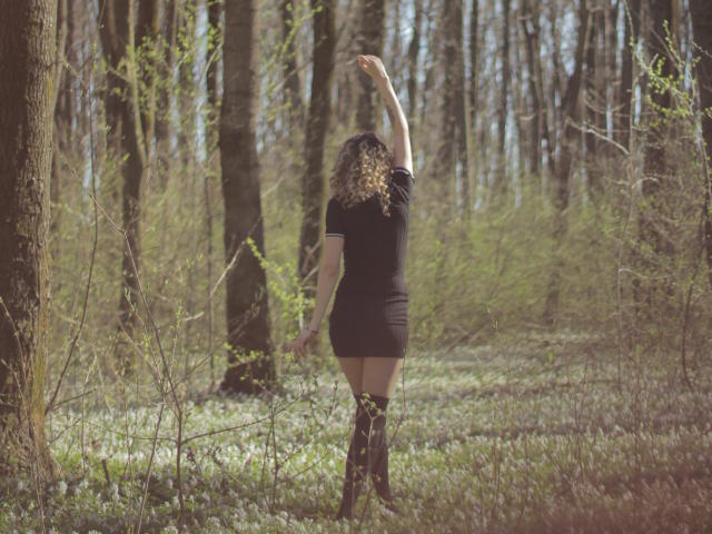 PoxyVibe stands in a forest with arms raised, wearing a black short dress and thigh-high socks, back to camera, curly hair, small ass, tree trunks in background.