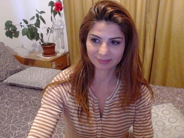 Sexydollhotx con capelli castani, camicia a righe, seduta sul letto, che guarda la fotocamera, sorridendo, con piccoli capezzoli rosa visibili, tende gialle sullo sfondo, pianta in vaso su un comodino di legno..