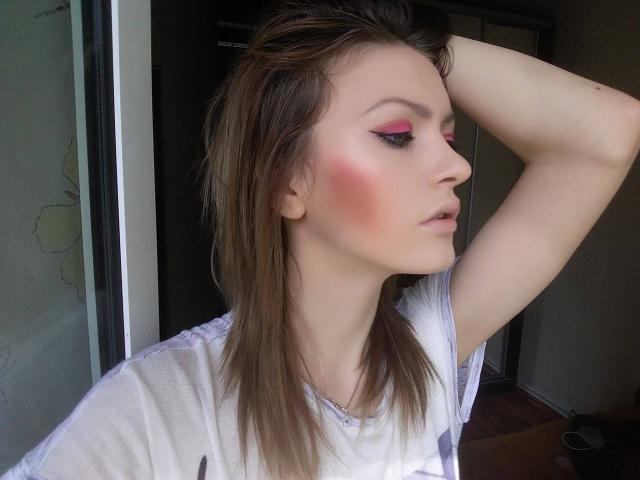 BlossomPussy, avec de l'ombre à paupières rose, un blush rosé et des cheveux bruns, levant son bras droit, portant une chemise blanche, regardant sur le côté avec les lèvres légèrement entrouvertes..
