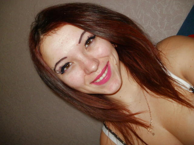 AnastasiyaXL with red-brown hair, smiling with pink lipstick, wearing a white tank top, showing her tits, and a gold necklace, looking at the camera.