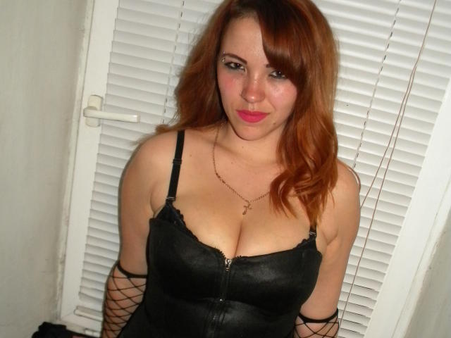 AnastasiyaXL with red hair, black lace-up bustier, and fishnet sleeves, showing her tits, pink lipstick, and a slight smile, standing in front of white blinds.