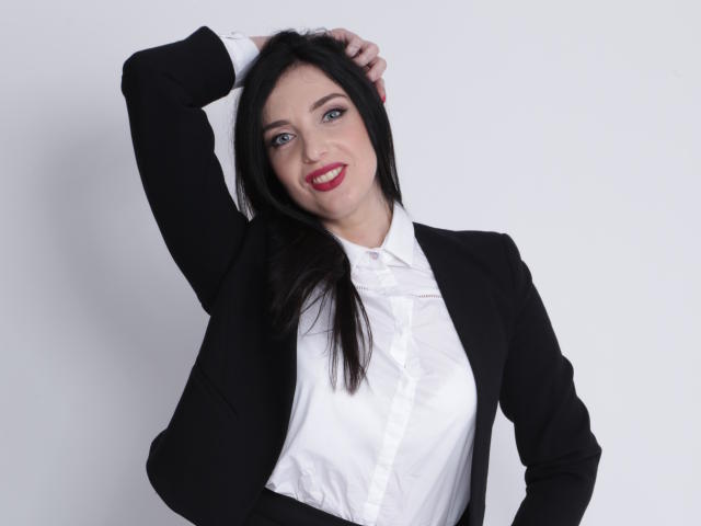 KatieFrenchie with black hair, red lipstick, white shirt, and black suit, hand in hair, smiling, showing her tits, standing against a white background.