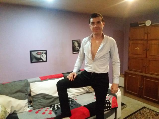 Bobandchris69, shirt unbuttoned, showing chest, black pants, sitting on bed with red, white, and gray blanket, wooden cabinet in background, looking at camera, slightly smiling.