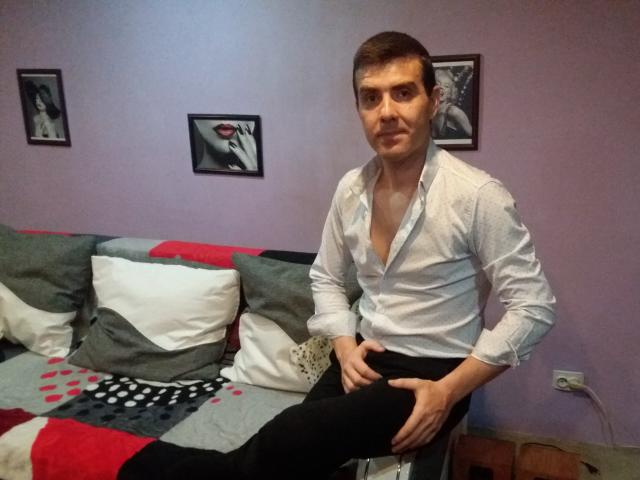 Bobandchris69 è un ragazzo con i capelli castano corti, che indossa una camicia bianca leggermente sbottonata, seduto su una sedia, pantaloni neri, mani sulle ginocchia, parete viola, foto in bianco e nero..