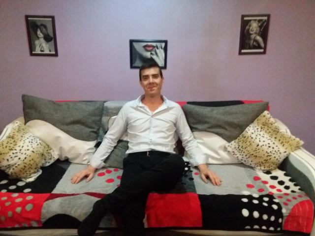 Bobandchris69 is a dude with short dark hair, wearing a white shirt and black pants, sitting on a colorful, patterned couch with gray and white pillows.