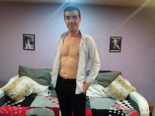 Bobandchris69, torse nu, portant un pantalon noir, les mains dans les poches, debout devant un canapé avec des coussins à motifs, dans une pièce aux murs violets avec des photos encadrées en noir et blanc..