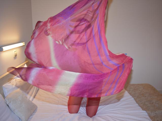 GinnyGold è inginocchiata su un letto, tenendo in mano una sciarpa trasparente rosa e viola che le copre il seno e i genitali. Si vedono le sue ginocchia e il letto ha lenzuola bianche..