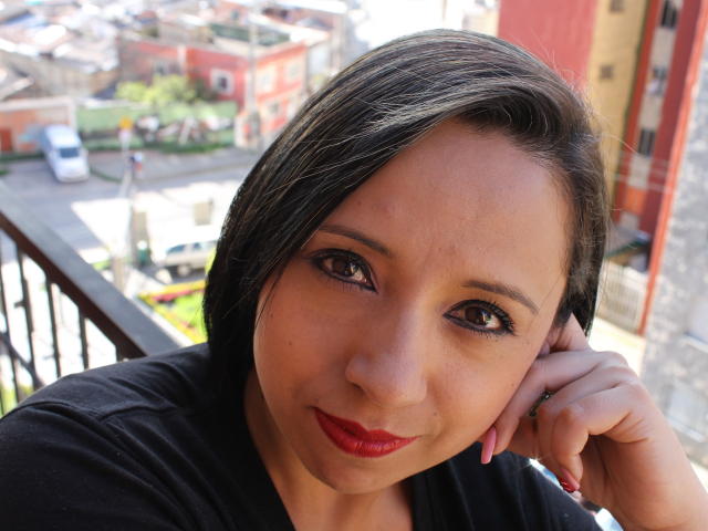 BellaLolita has medium brown skin, black shoulder-length hair, and red lipstick. She's smiling with her hand on her cheek, wearing a black shirt, and has brown eyes.