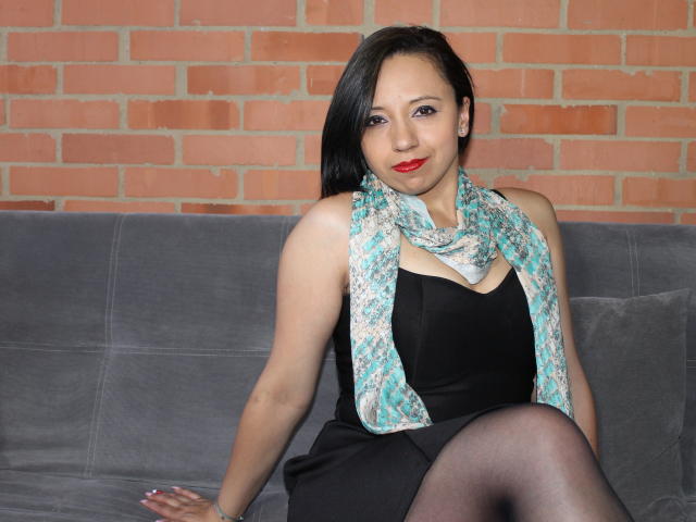 BellaLolita with black hair, red lipstick, black dress, teal scarf, and black tights, sitting on grey couch, showing her tits and ass, against red brick wall.