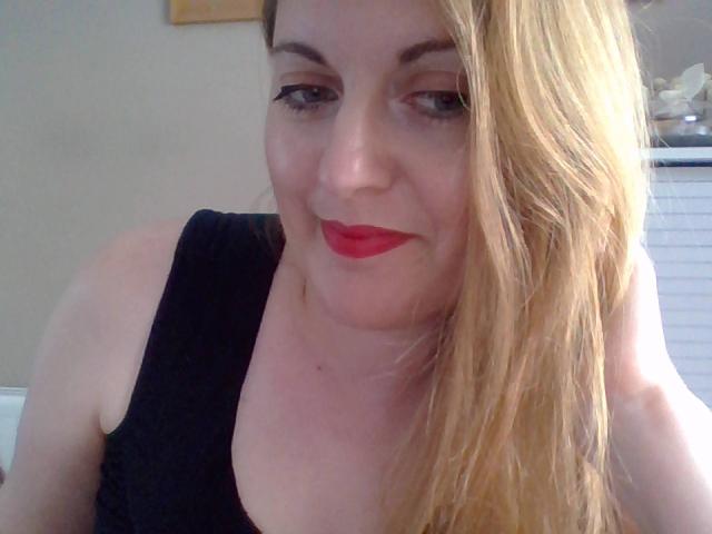 FrenchLea, con i capelli biondi, il rossetto rosso, la canottiera nera e un leggero sorriso, mostra il seno e i glutei nella foto. L'eyeliner è visibile e sembra carina..