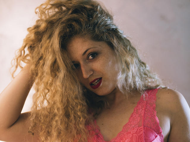 SassyKate with curly blonde hair, red lipstick, and pink lace bra, looking seductively at the camera, hand in hair, smiling. Boobs visible, freckles, sexy, confident.