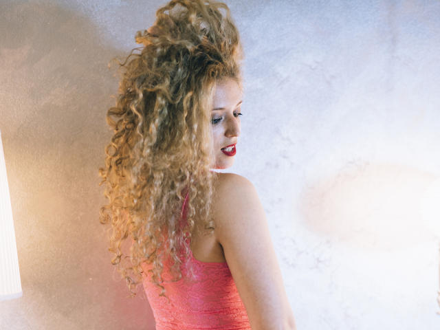 SassyKate with long, curly blonde hair, wearing a pink lace dress, looking over her shoulder, smiling with red lipstick, soft light on the left, white textured wall.