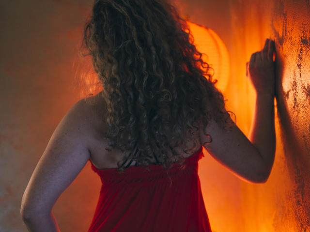 SassyKate with curly brown hair, back to camera, wearing a red sheer dress, touches a wall, glowing orange light from behind. Her ass is slightly visible.