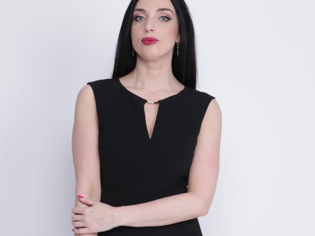 KatieFrenchie with long black hair, red lipstick, and black sleeveless dress, arms crossed, showing her small tits, pale skin, and pink nails, standing against a plain white wall.