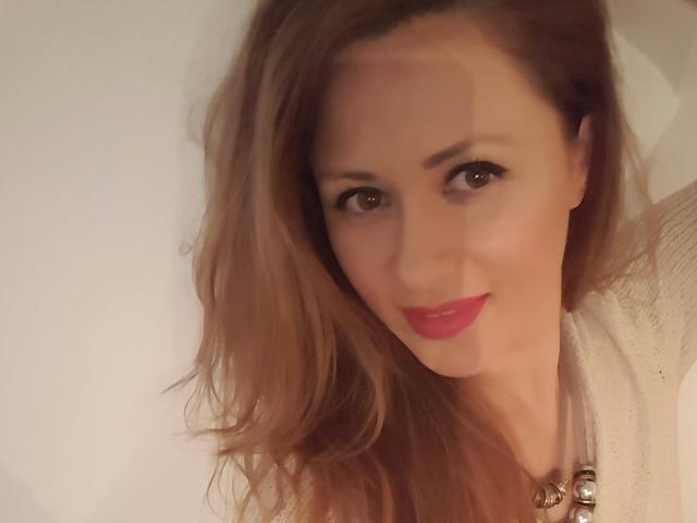 ToiSecret hat welliges, braunes Haar, roten Lippenstift und ein leichtes Lächeln und trägt einen weißen Pullover mit einer Halskette. Ihre braunen Augen sind sanft fokussiert..