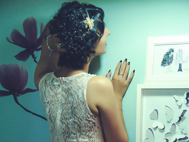 AmethystCharm with curly dark hair, white lace dress, flower hairpin, black nails, standing, back to camera, hand on wall, looking at framed art, soft green wall.