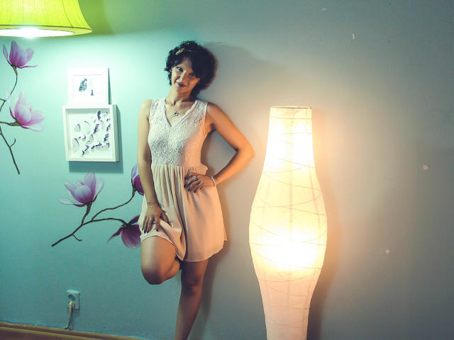AmethystCharm with short curly hair, wearing a white lace dress, standing against a wall, leg raised, next to a tall, glowing lamp, smiling, looking right.