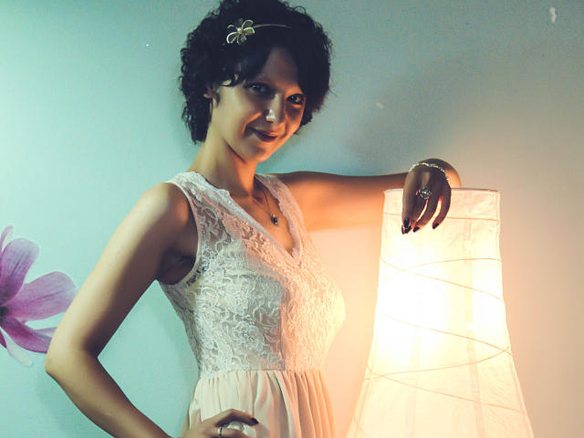 AmethystCharm with curly black hair, wearing a white lace dress, stands next to a lit lamp, smiling, with a green hairpin and ring, showing her tits and ass.