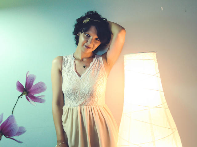 AmethystCharm with short curly hair, wearing a white lace dress and pink skirt, standing by a lit paper lantern, smiling with one arm raised, showing her tits.