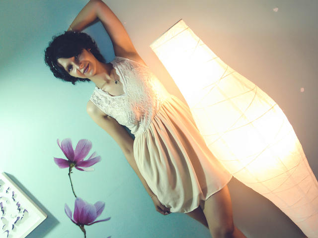 AmethystCharm with curly dark hair, wearing a white lace sleeveless dress and a necklace, standing with arm raised, next to a lit paper lantern and purple flowers.